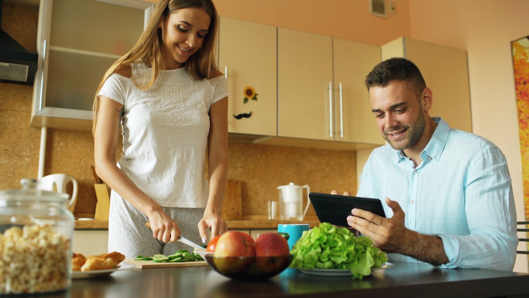 An attractive couple is chatting in the kitchen early in the morning. The handsome man is using a tablet while his girlfriend prepares breakfast at home