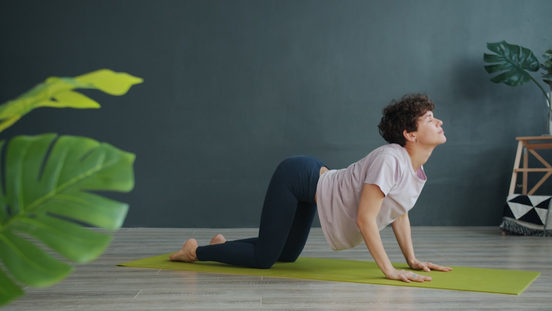 A cute female yogi is doing spinal exercises indoors at a fitness center; she enjoys yoga by working out alone. The serious woman is wearing modern sportswear and exercises barefoot.