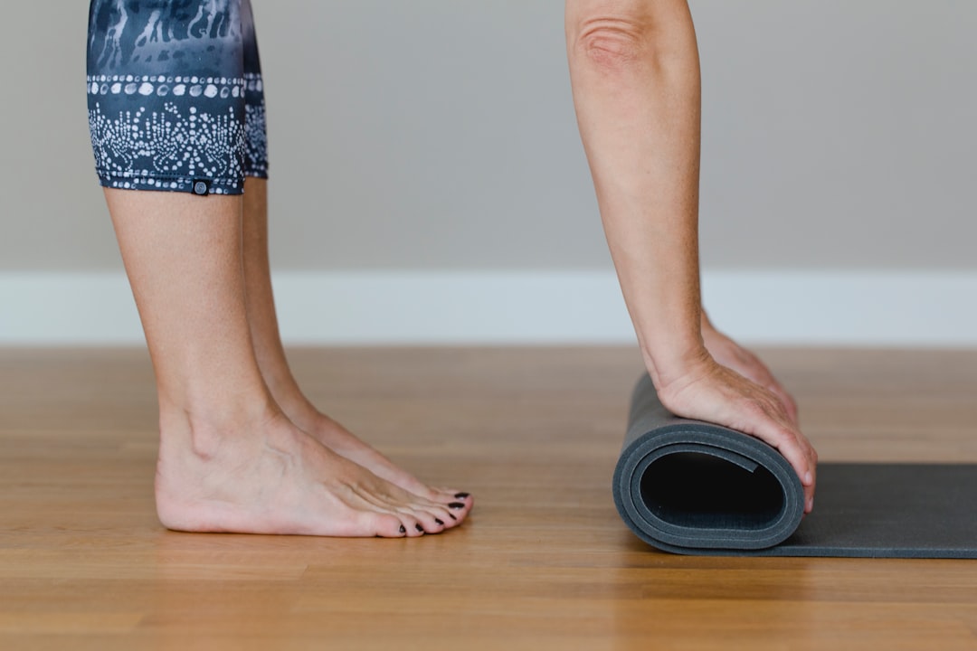 A woman is rolling up her yoga mat in a yoga studio. While doing this, she stands upright and keeps her feet firmly planted on the ground. The yoga mat is blue, and it’s the end of that day’s yoga class.