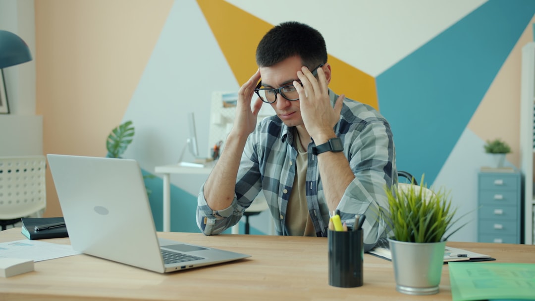 A stressed man with glasses suffers from an overwork-related headache while working on a laptop in the office; he touches his head and rubs his temples. Work and health concept.