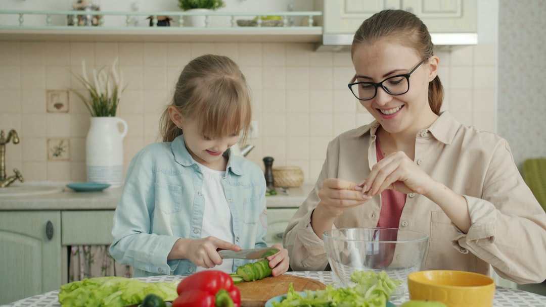 A helpful girl is in the kitchen at home, slicing cucumber and making a vegetable salad, while her young and loving mother shows her affection by touching her hair.