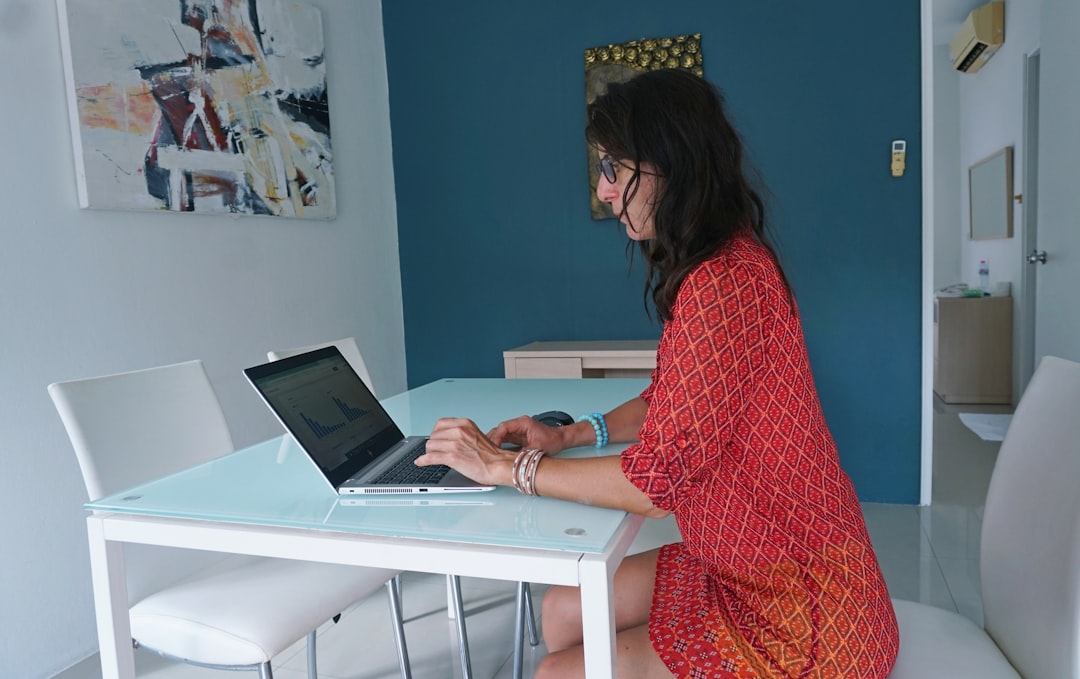 A freelance businesswoman in a red dress works from home, using web-based and mobile technologies to produce instantly updatable and shareable content for training programs. The design of her apartment is minimal and comfortable, helping her focus on tasks.