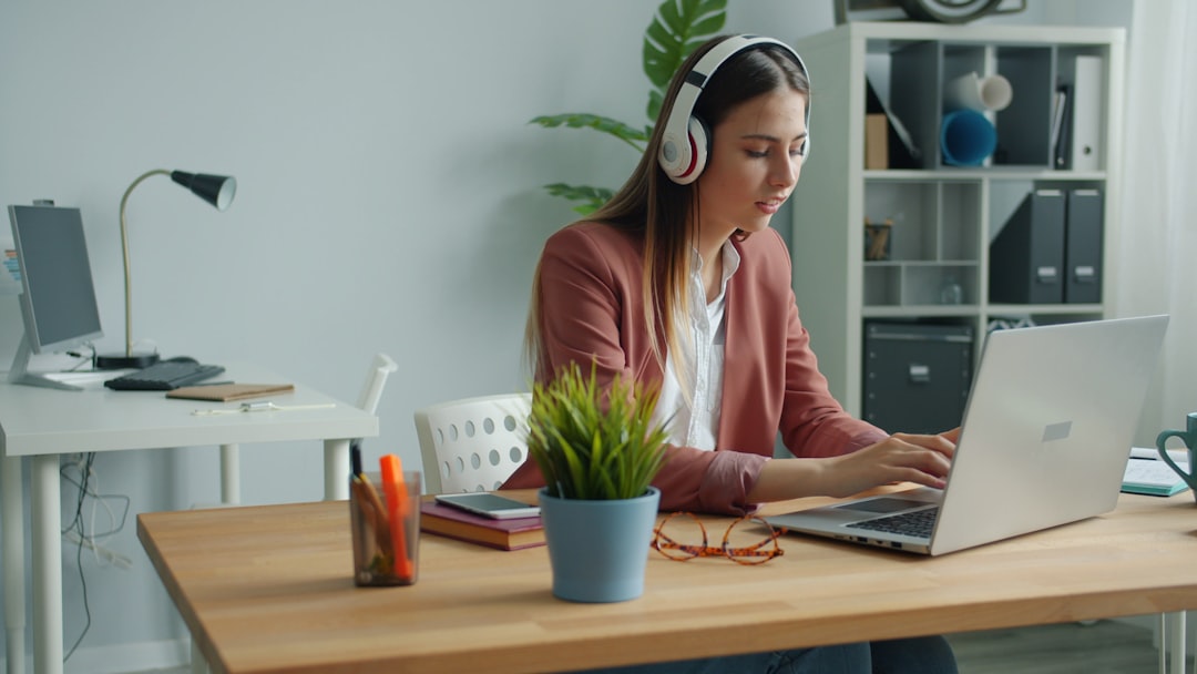 A cheerful manager listens to music with wireless headphones, sings, and types using a computer at work; sitting alone at the desk. People, business, and entertainment concept.