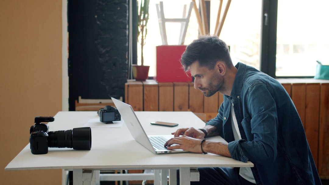 A serious bearded photographer edits photos after a shoot in the studio using a laptop; typing while working alone, with a camera visible on the desk. People and business concept.