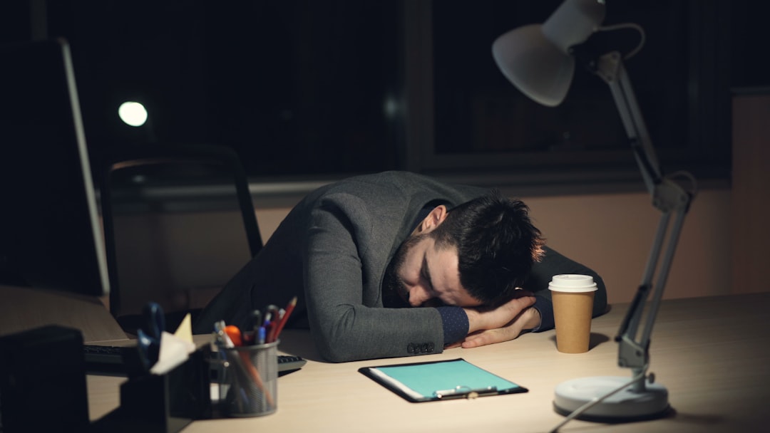 A tired man in a formal suit sleeping on his office desk late at night after an intense workday. An open laptop, takeaway coffee, and a lamp are seen on the desk.