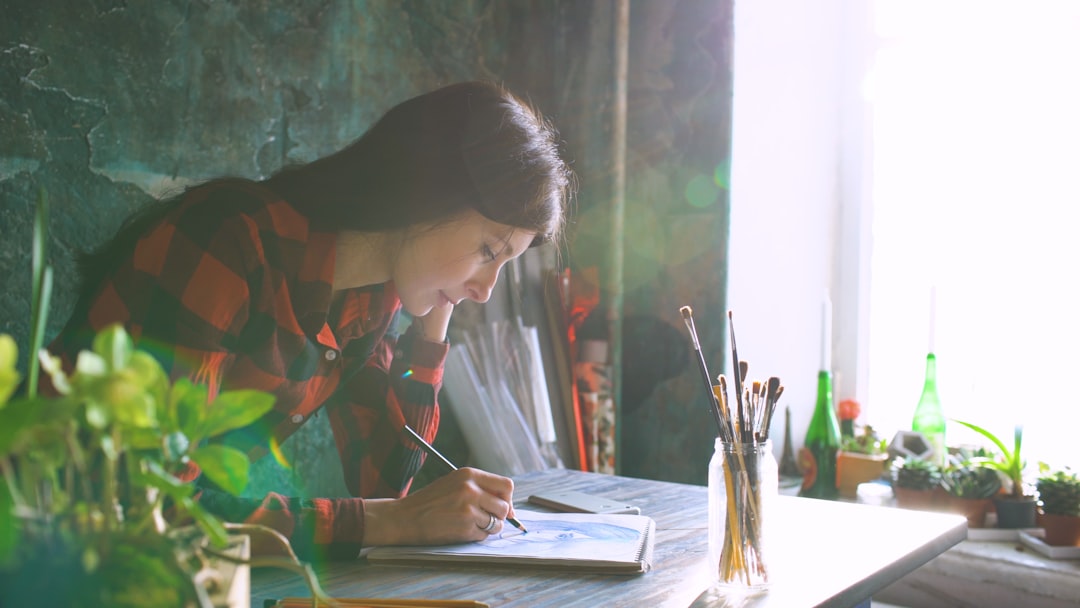 Young female artist sketching with a pencil in a paper notebook indoors in a studio. Speckles of sunlight