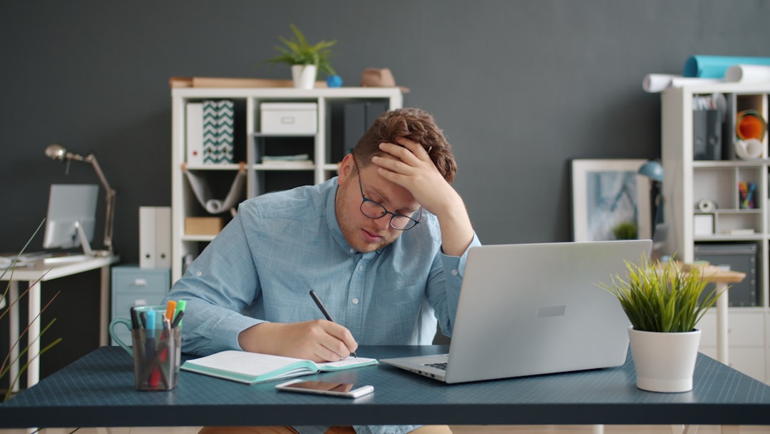 The tired young man is typing and working at a computer indoors at the workplace; he is experiencing a headache, touching his face and head. The concept of workaholism, work, and human health.