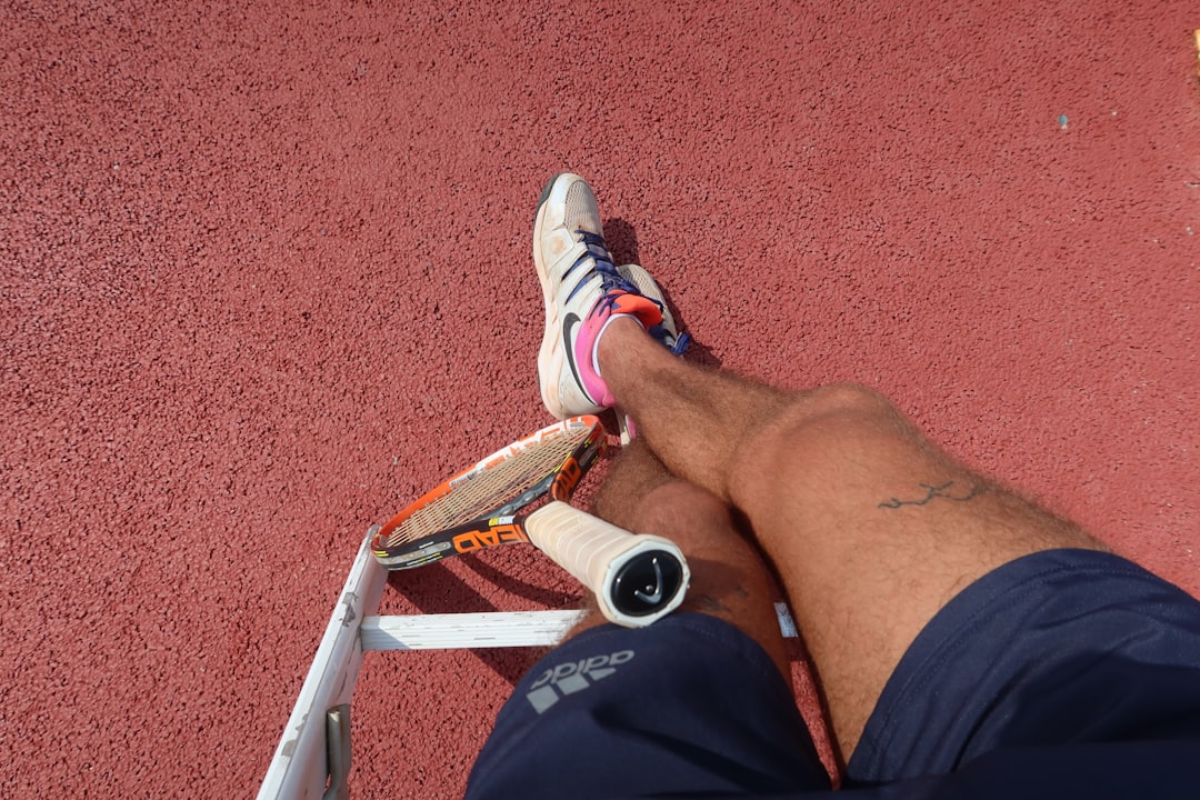 A close-up of a tennis player sitting cross-legged on a red hard court; resting with a HEAD tennis racket leaning against a low white chair, wearing Adidas shorts and Nike athletic shoes, and a small ankle tattoo is visible.